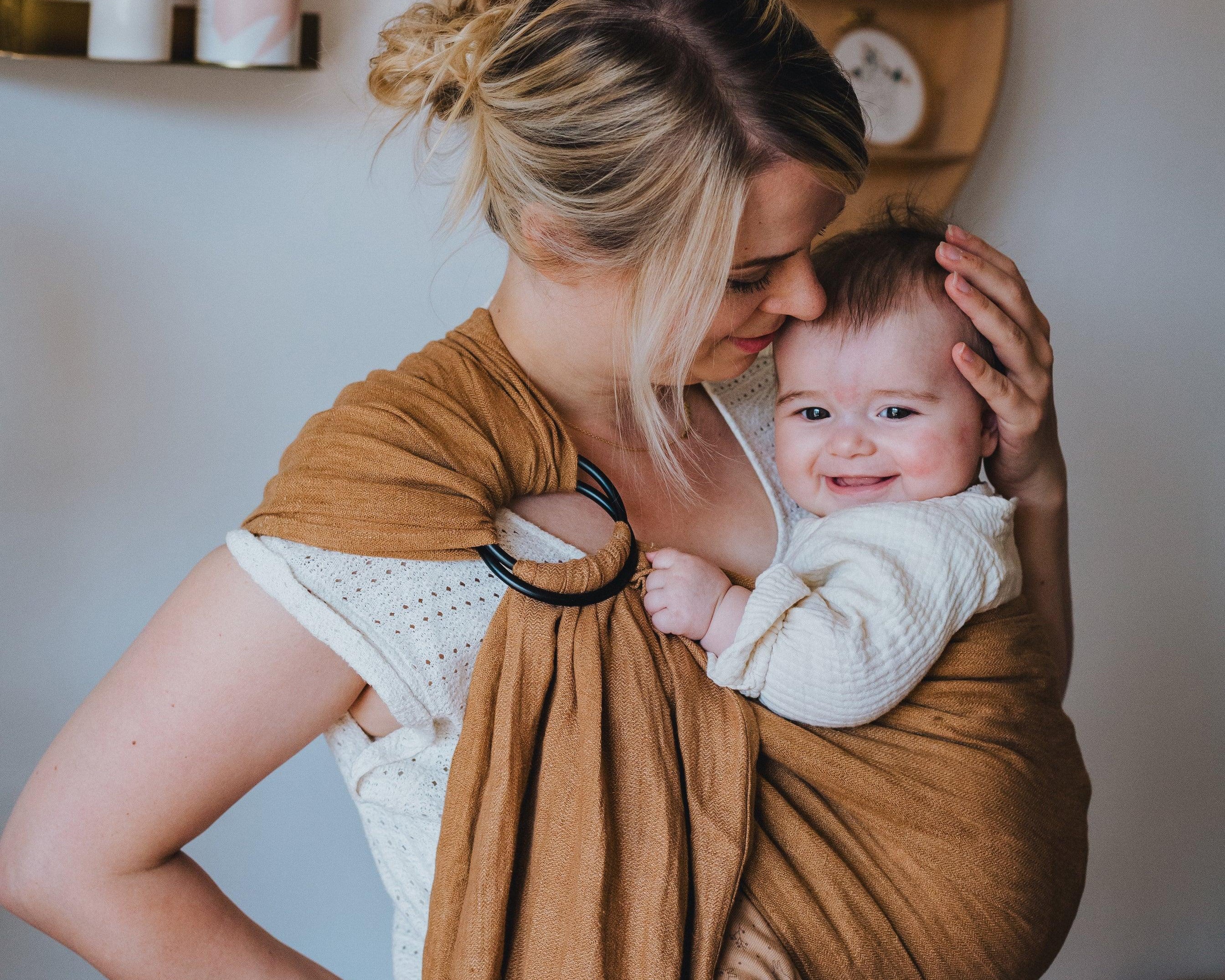 maman qui caresse son bébé souriant dans un ring sling bébé marron 