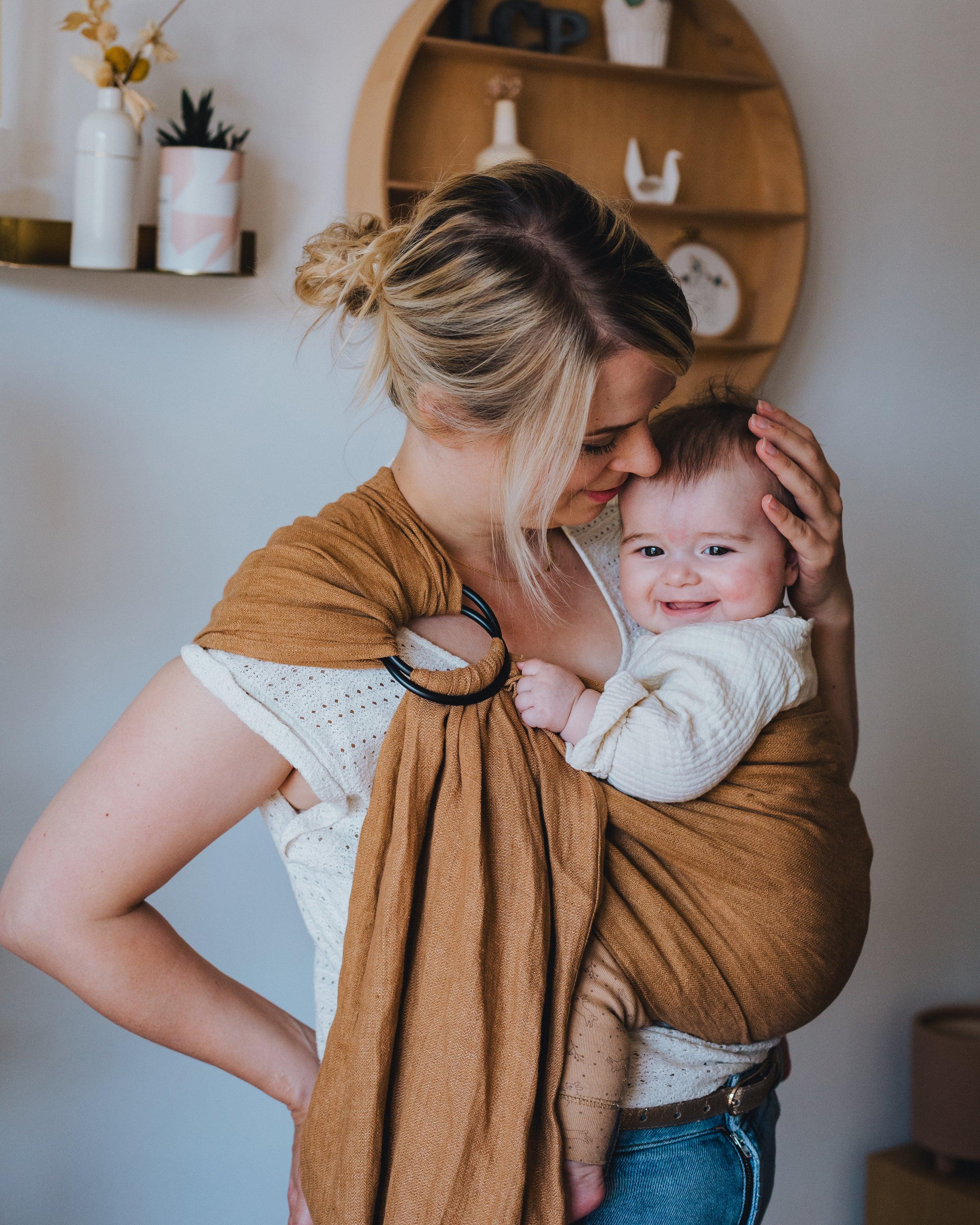 maman qui caresse son bébé souriant dans un ring sling bébé marron 