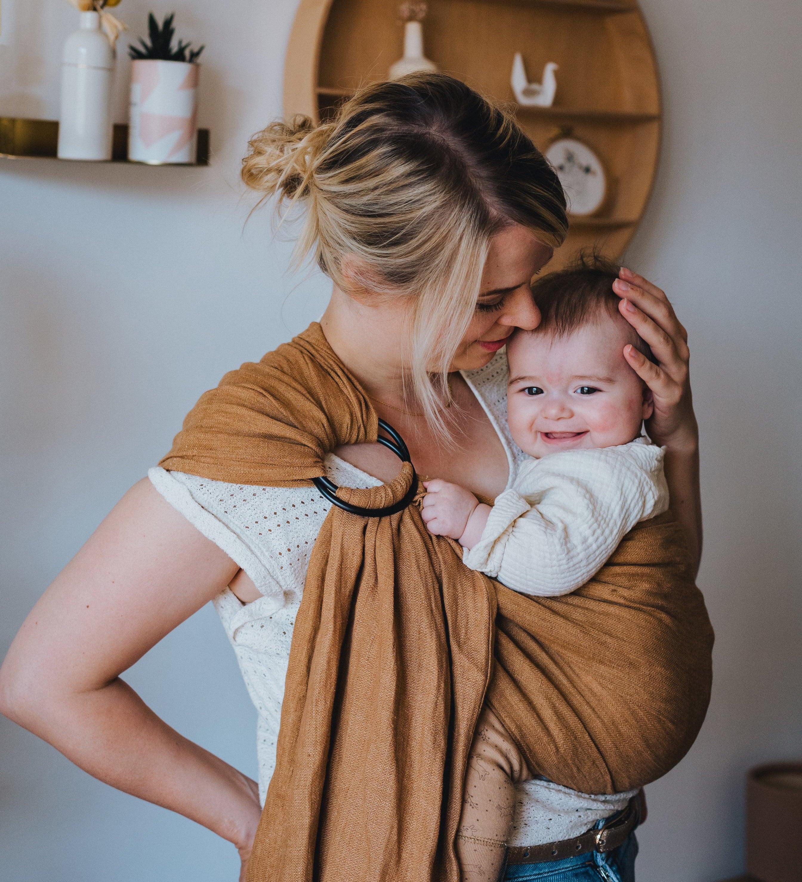 maman qui caresse son bébé souriant dans un ring sling bébé marron 