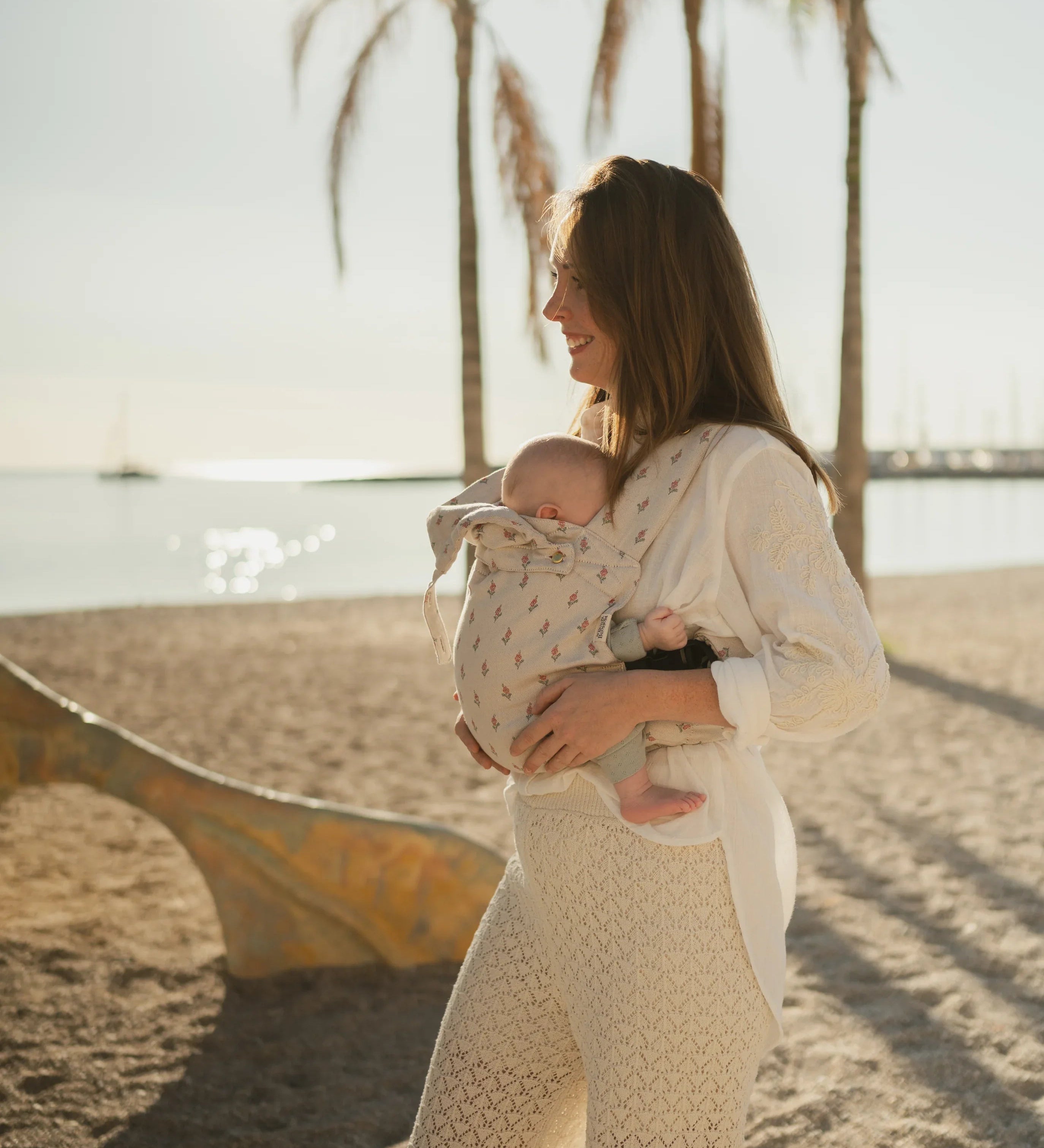 Maman portant son bébé en porte-bébé physiologique Bud & Blossom sur une plage en été