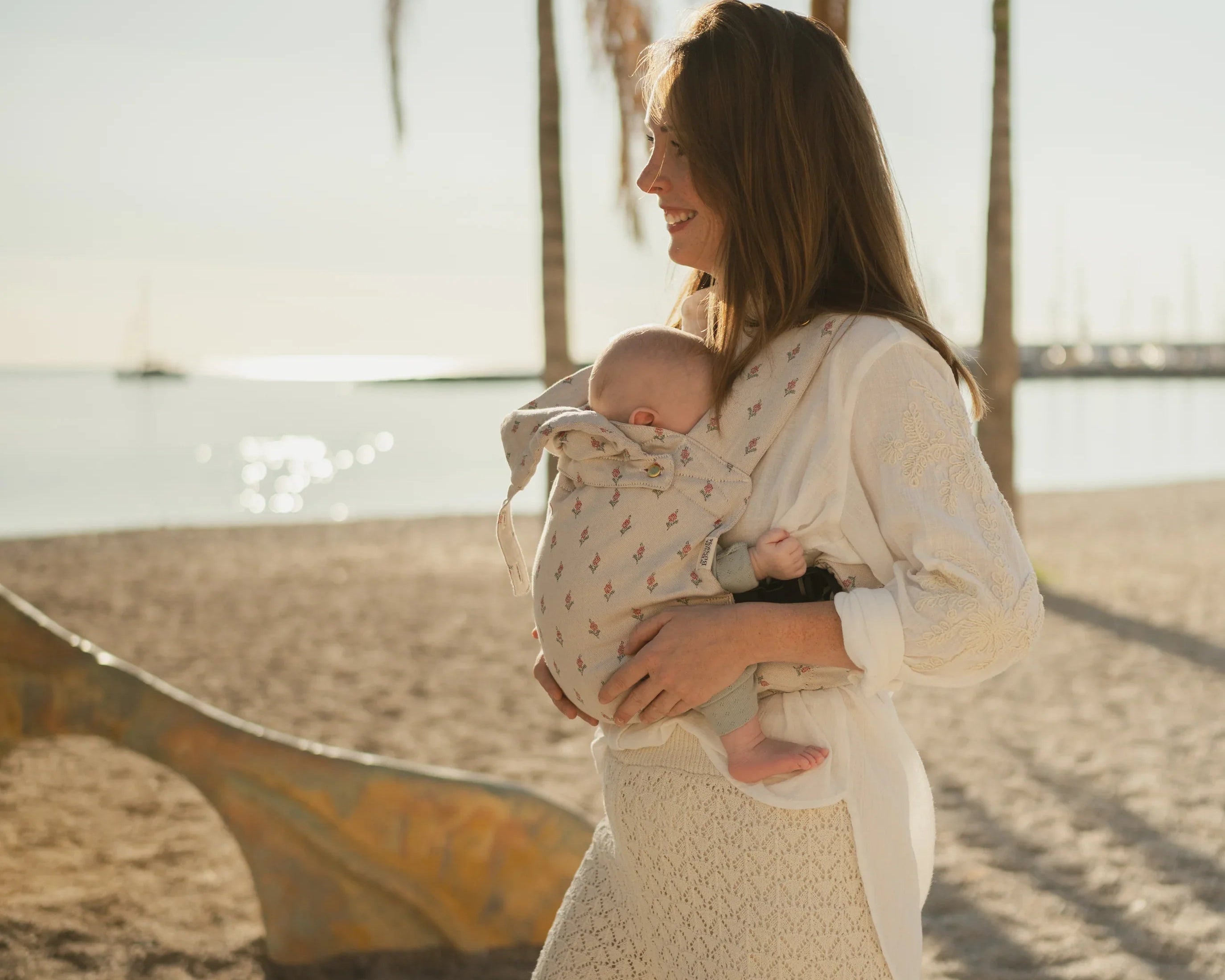 Maman portant son bébé en porte-bébé physiologique Bud & Blossom sur une plage en été
