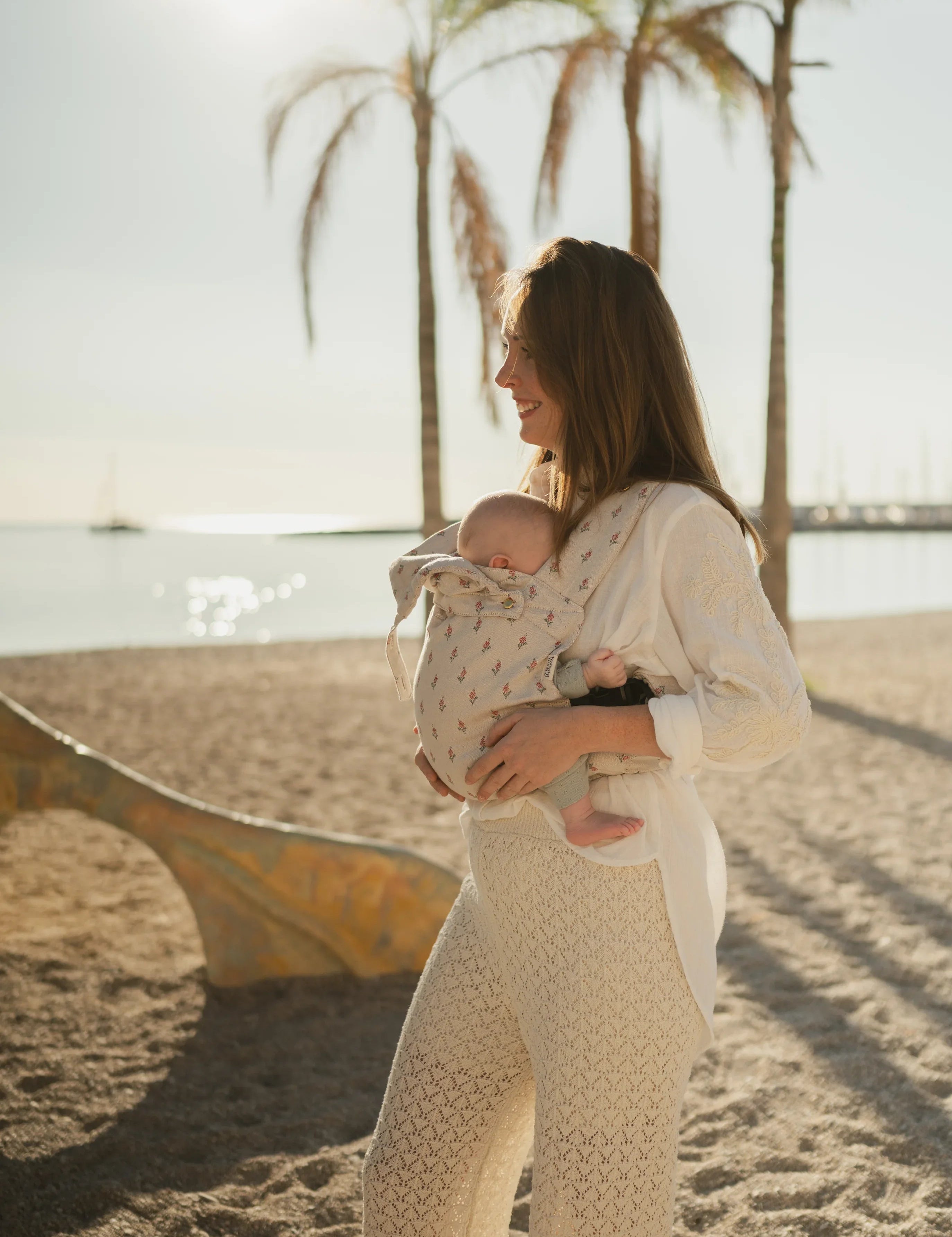 Maman portant son bébé en porte-bébé physiologique Bud & Blossom sur une plage en été