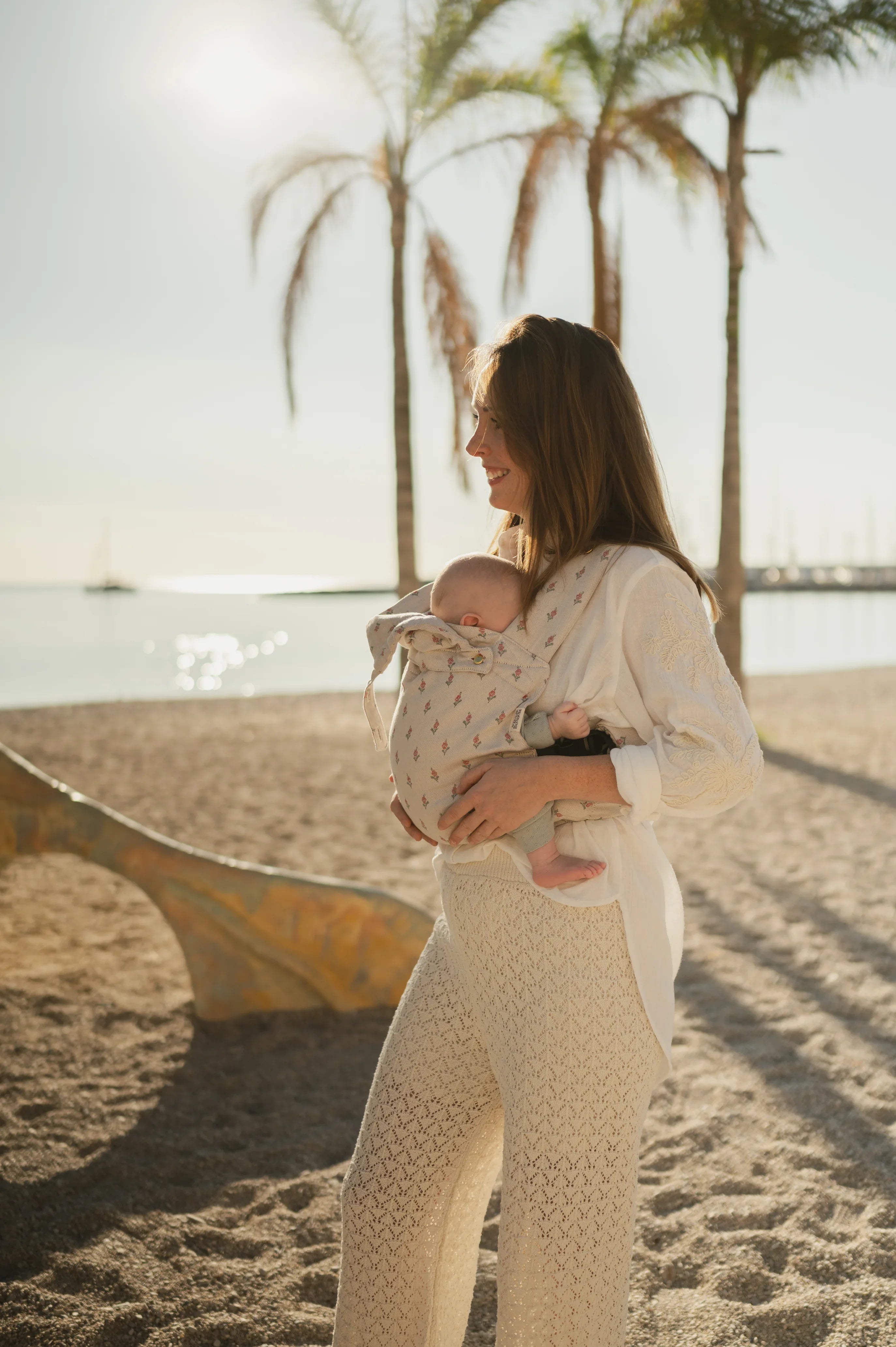 Maman portant son bébé en porte-bébé physiologique Bud & Blossom sur une plage en été