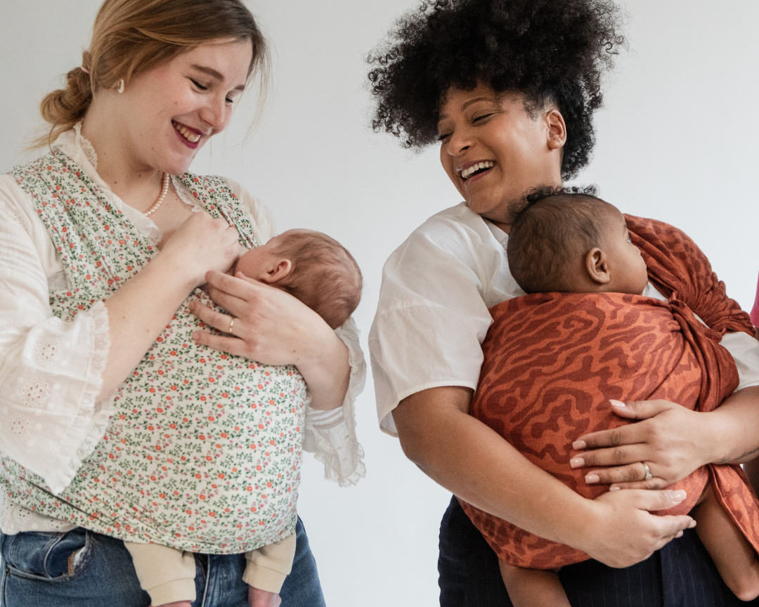 Deux femmes portent chacune un bébé dans un moyen de portage physiologique : l’une avec une écharpe extensible Wrap Prairie, l’autre avec un Sling en lin Curve terracotta.