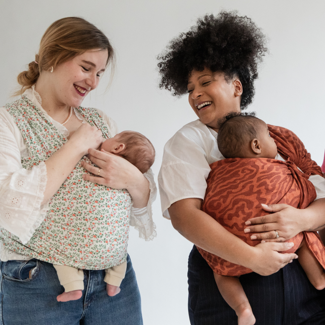 Deux femmes portent chacune un bébé dans un moyen de portage physiologique : l’une avec une écharpe extensible Wrap Prairie, l’autre avec un Sling en lin Curve terracotta.