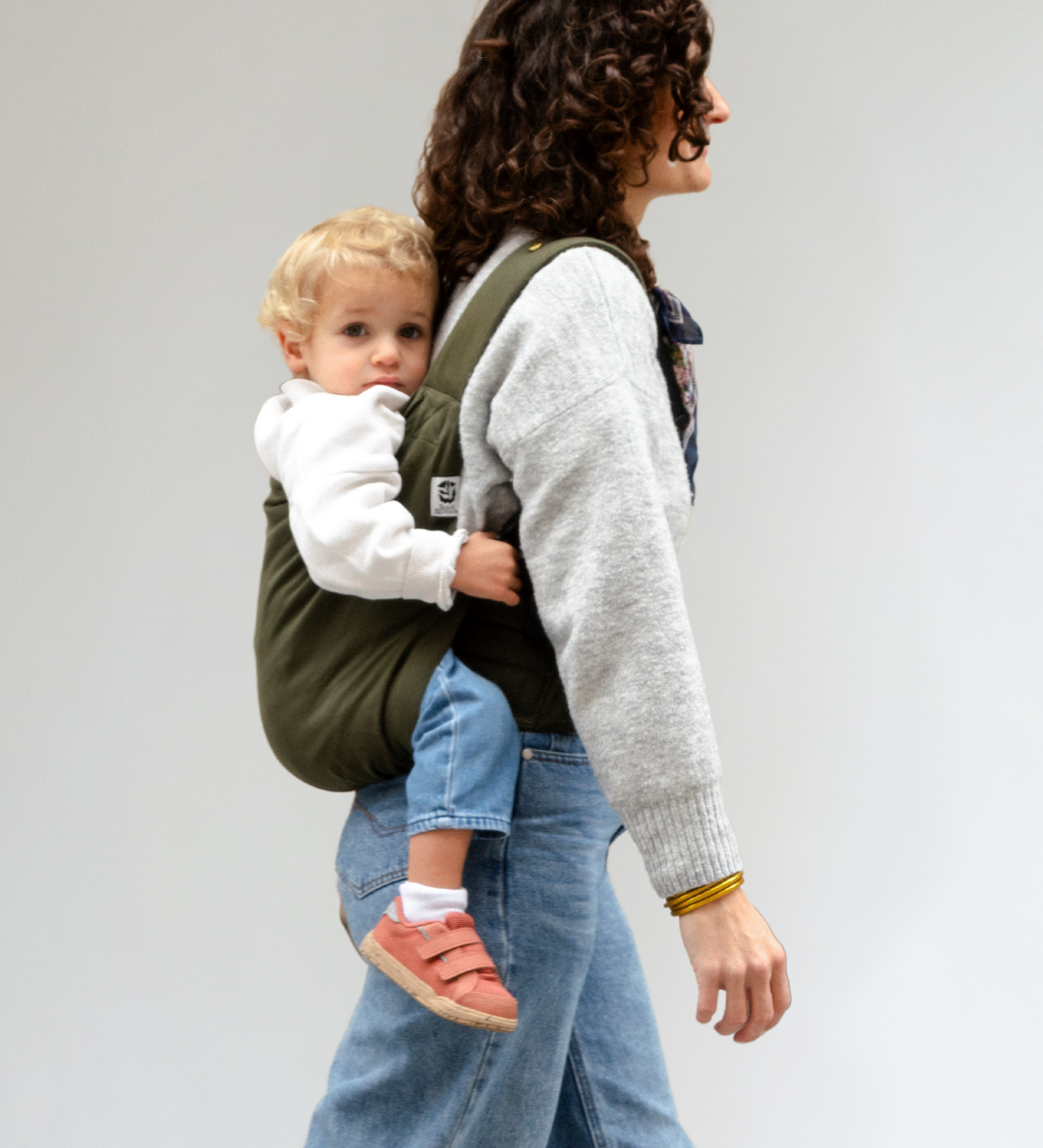 Maman portant son bambin sur le dos avec le porte-bébé préformé Exquis de Bud & Blossom, vue de côté en train de marcher.