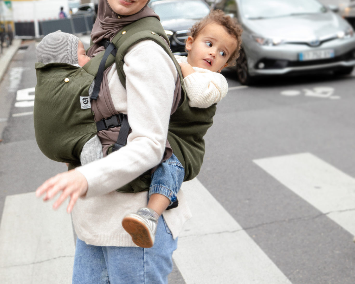Photo d’une maman portant ses deux enfants en porte-bébés dans une rue parisienne.