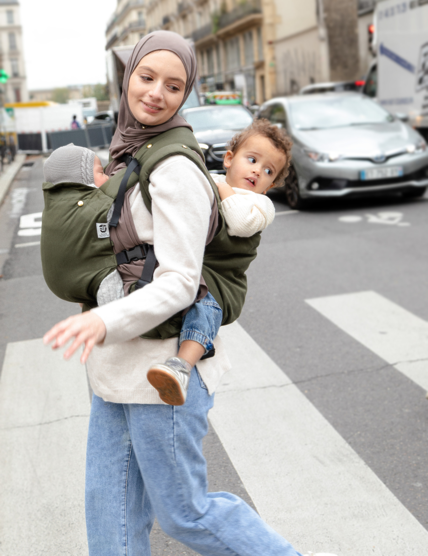 Photo d’une maman portant ses deux enfants en porte-bébés dans une rue parisienne.
