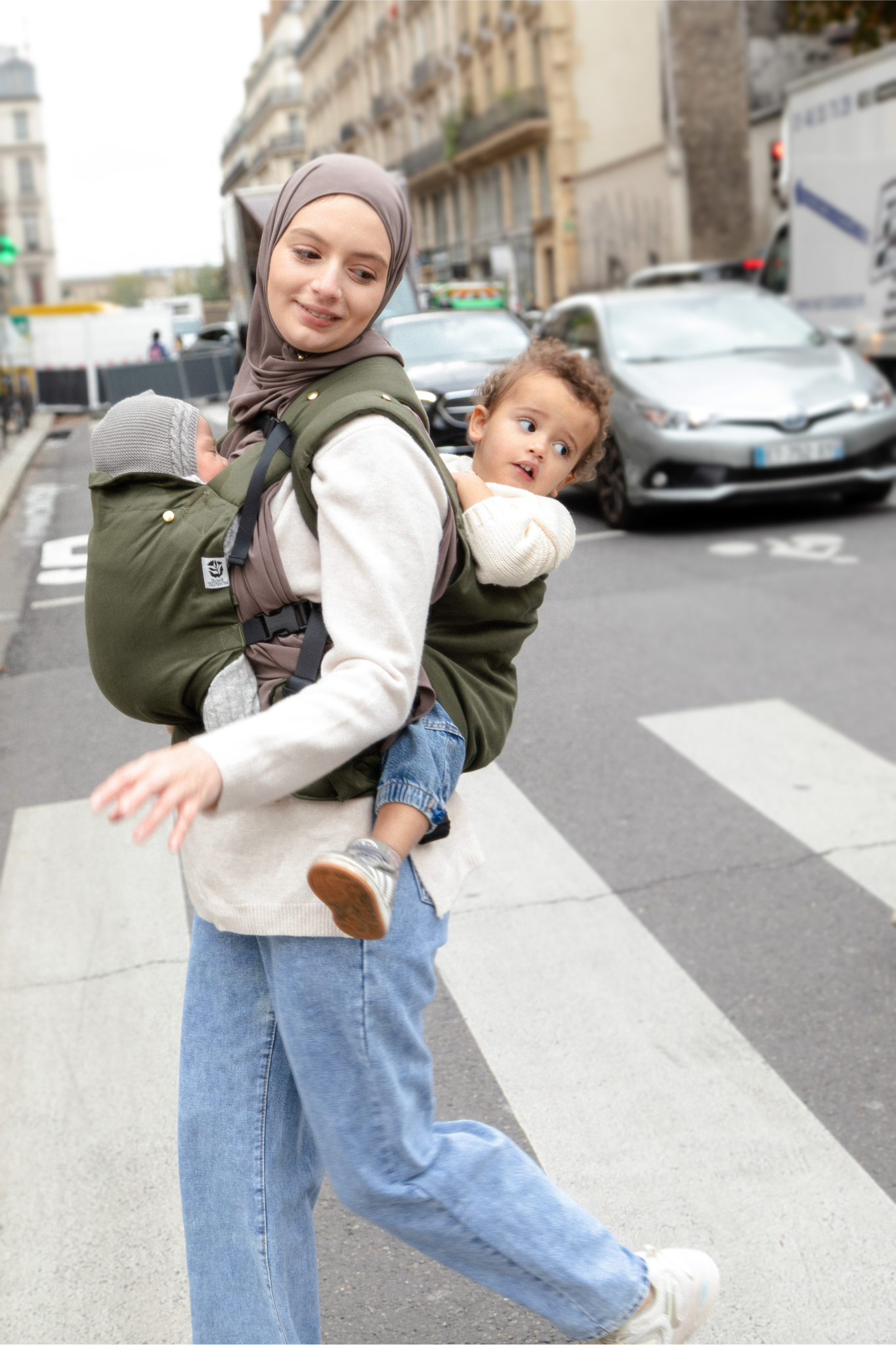 Photo d’une maman portant ses deux enfants en porte-bébés dans une rue parisienne.