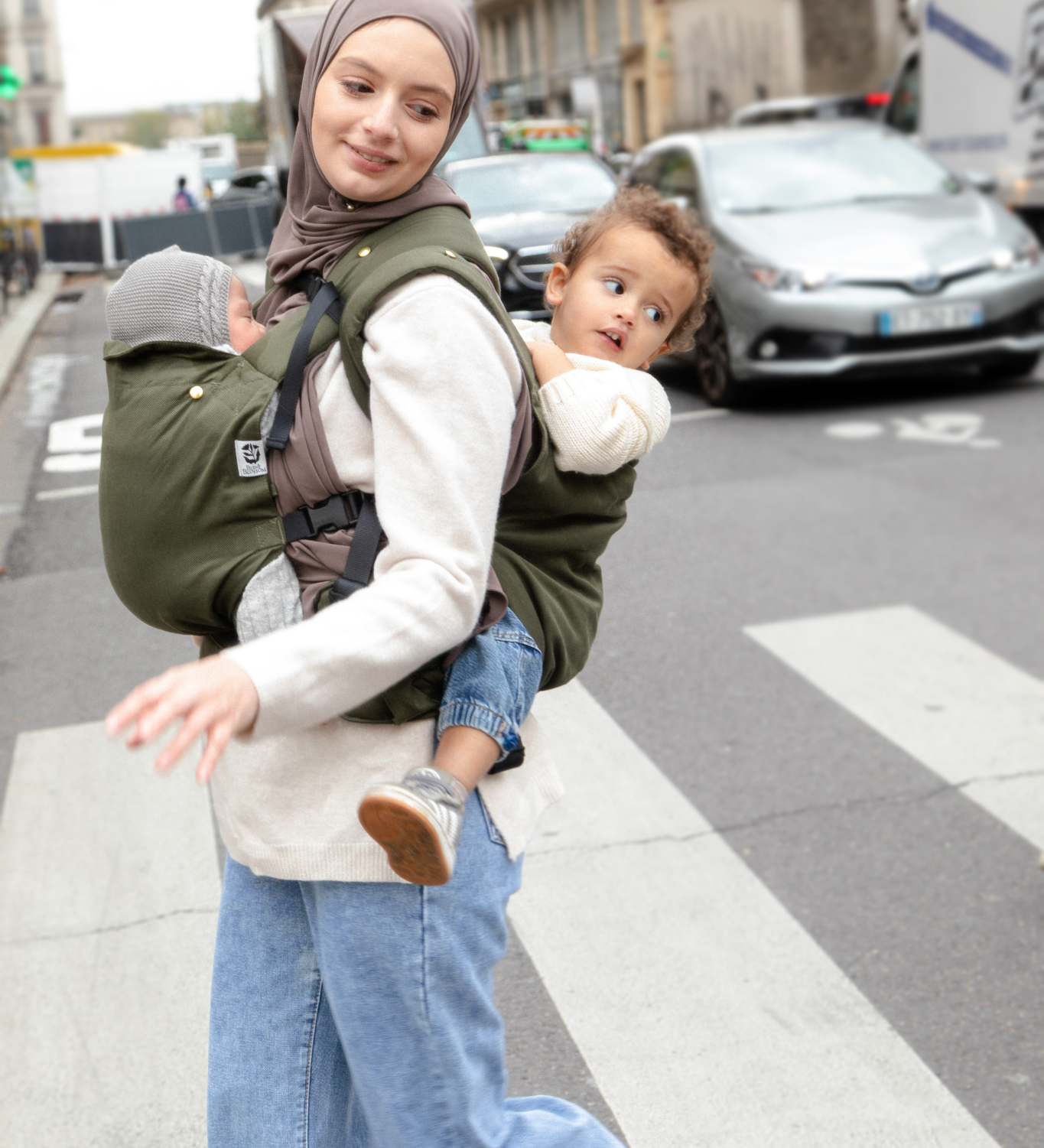 Photo d’une maman portant ses deux enfants en porte-bébés dans une rue parisienne.
