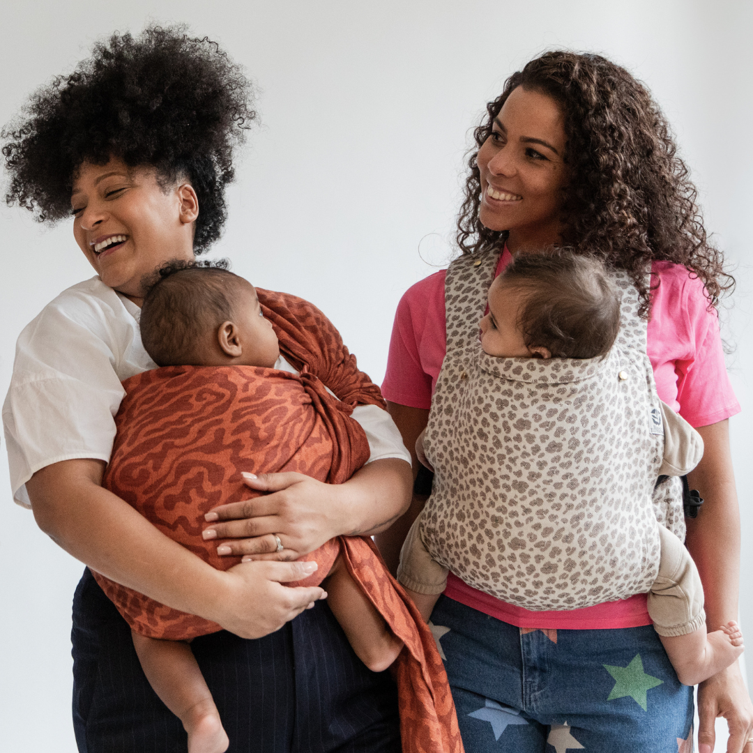 Deux mamans souriantes portant leur bébé de 6 mois, l'une avec le porte-bébé physiologique Exquis Leo, l'autre avec un Sling Curve