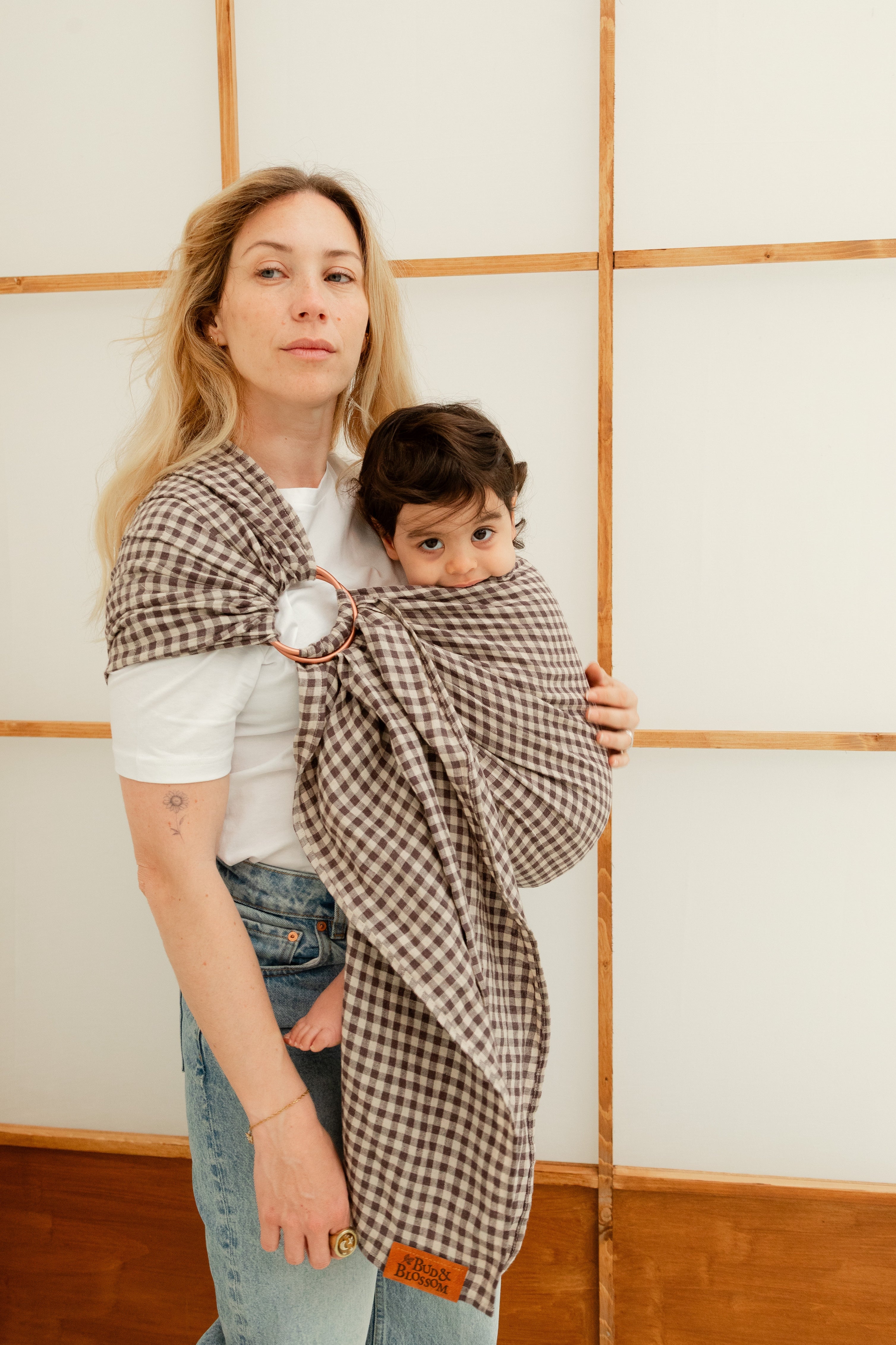 Maman portant sa fille de 24 mois en sling en lin, tandis que l’enfant met le tissu du sling à la bouche dans un moment complice.