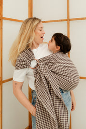 Maman portant sa fille de 24 mois dans un sling en lin, échangeant une grimace complice pleine de tendresse.