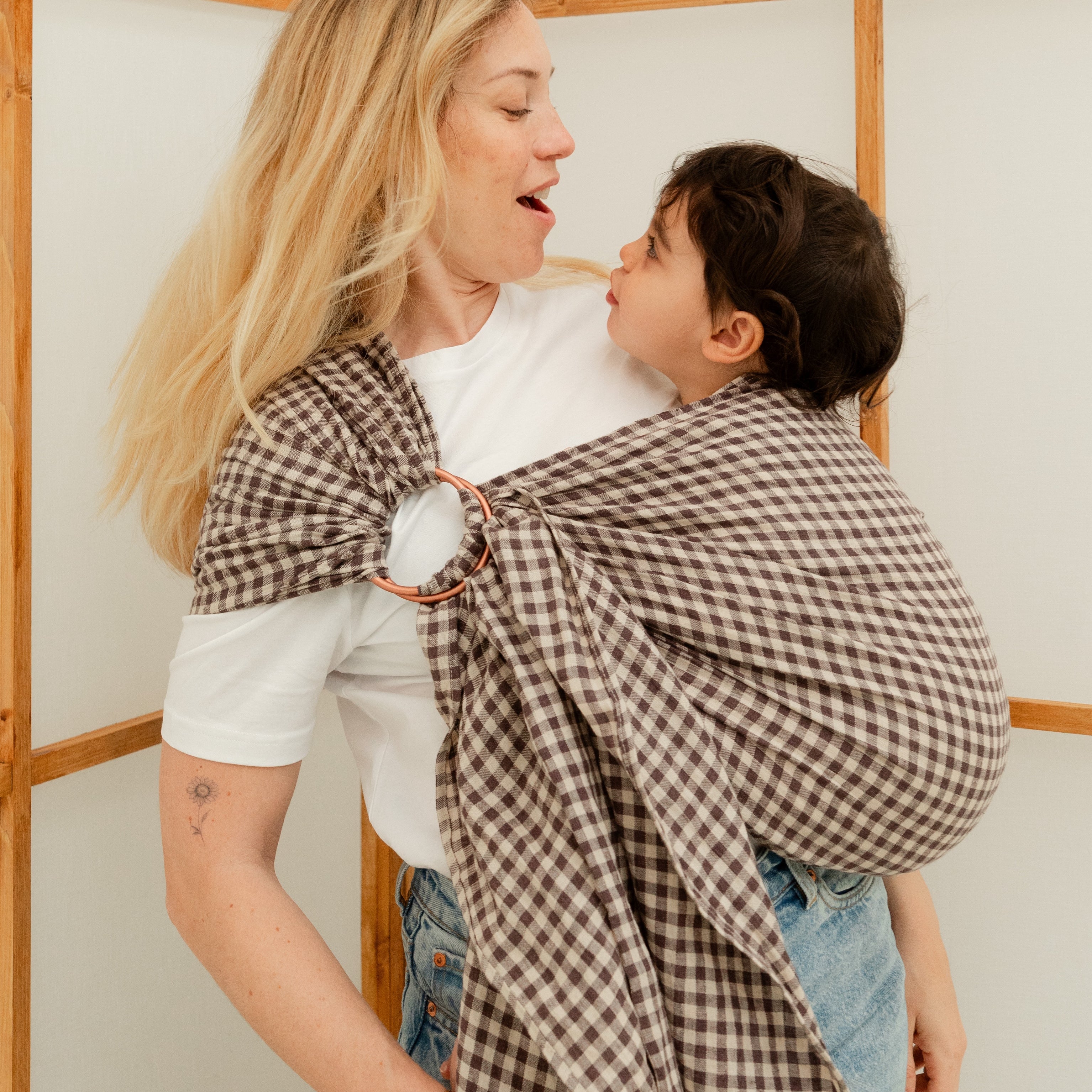 Maman portant sa fille de 24 mois dans un sling en lin, échangeant une grimace complice pleine de tendresse.