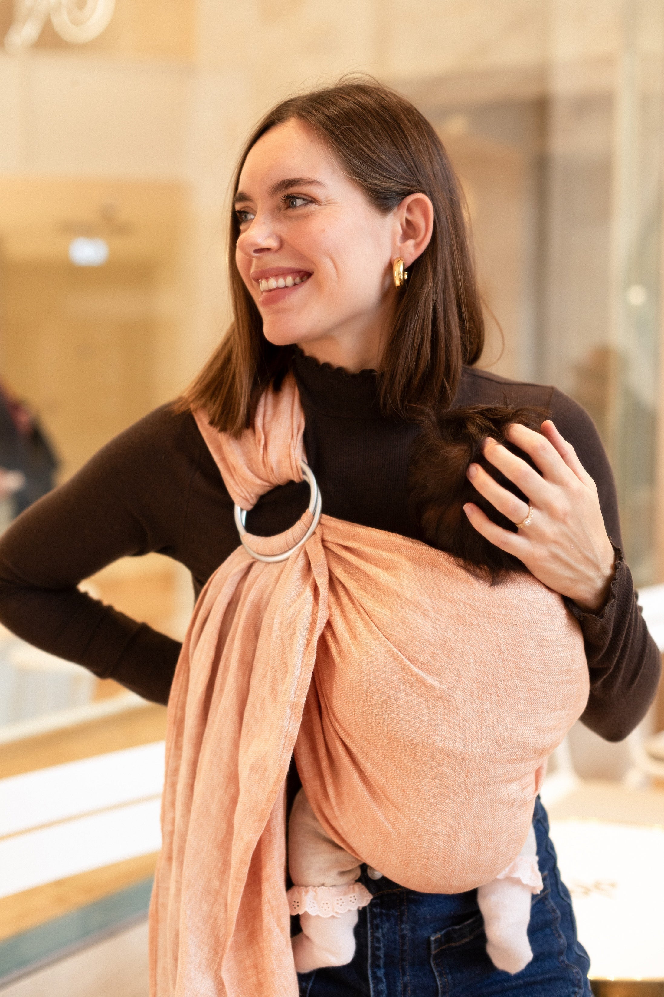 Sling en lin Paloma Bud &amp; Blossom coloris corail, porté par une maman dans un café avec son bébé de 3 mois confortablement installé.