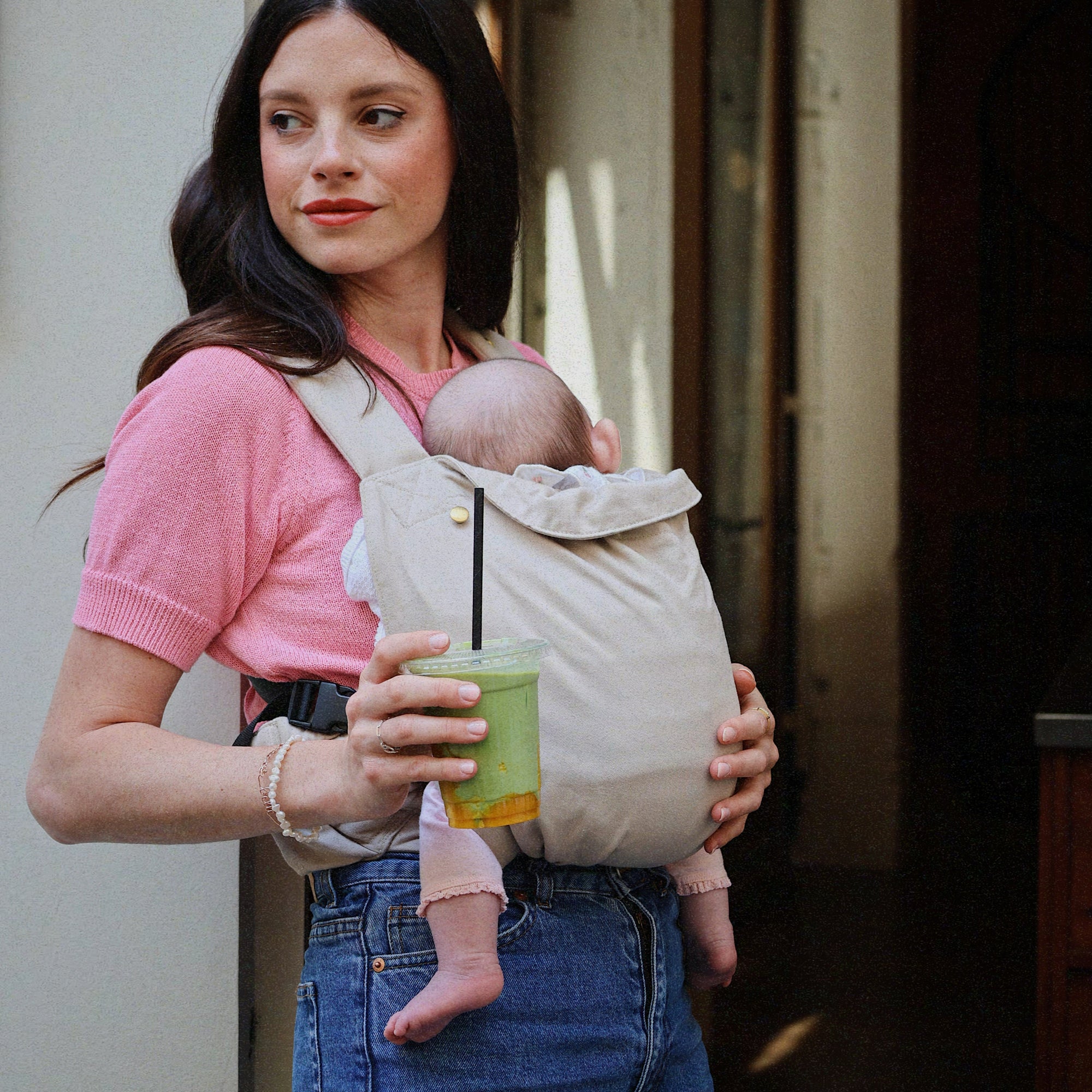 Maman brune portant le porte-bébé Exquis Bud &amp; Blossom coloris taupe, bébé blotti contre elle, tenant un ice matcha latté, appuyée contre un mur dans un café.